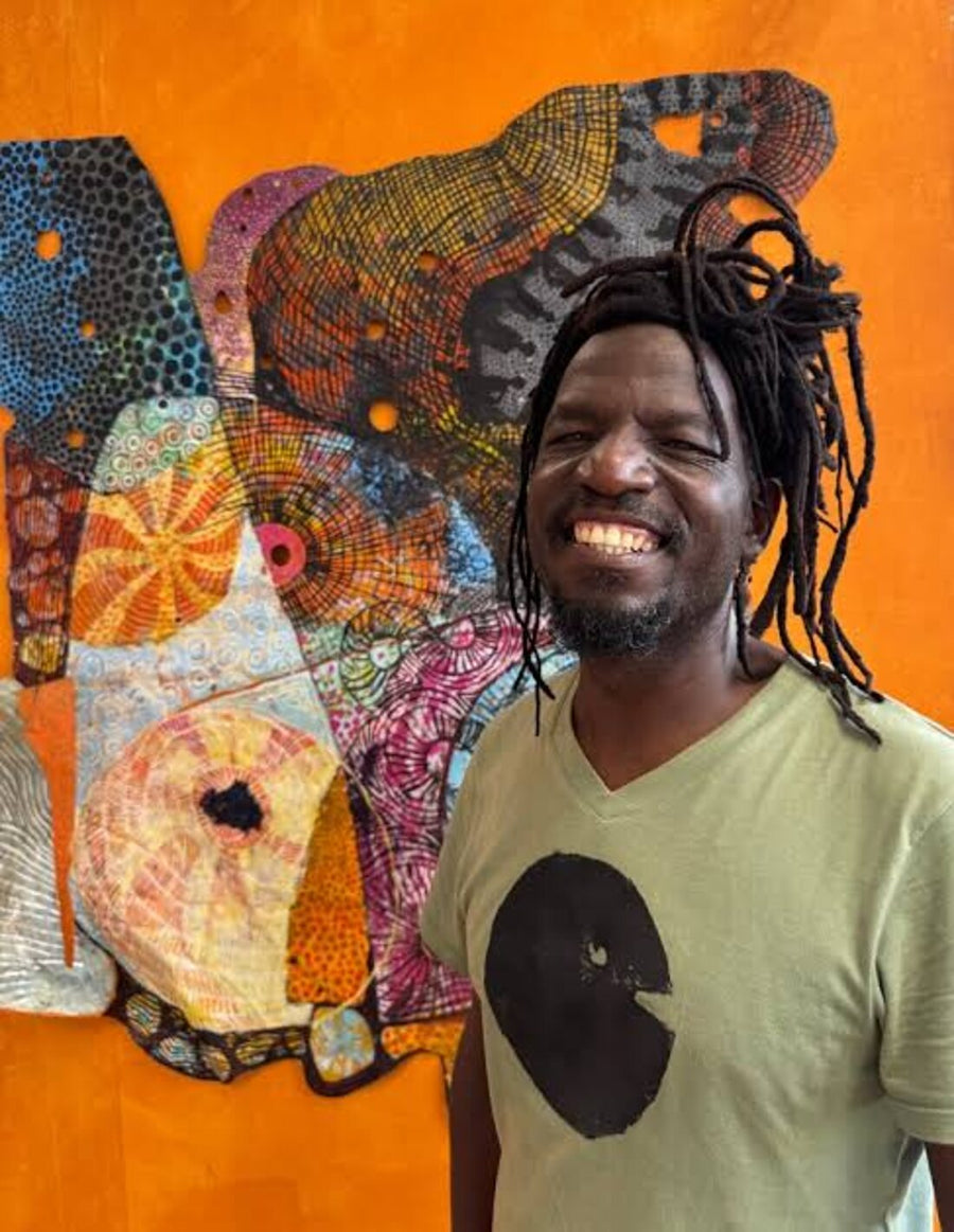 A smiling Jonathan stands in front of a bright orange wall featuring his colorful abstract mosaic artwork.